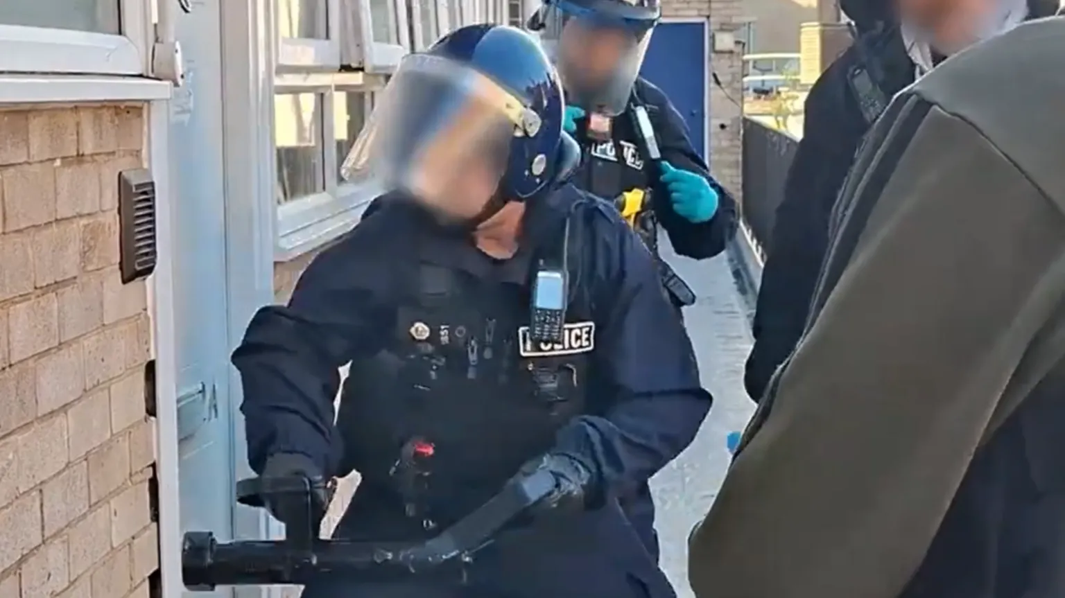 Norfolk Police Police officer in tactical gear breaking a door down using a battering ram tool. There are other officers nearby. Their faces have been blurred,