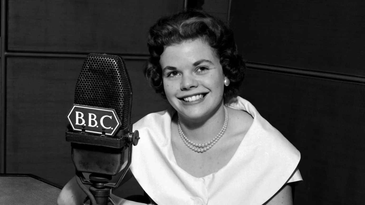 Black & white photo of a young Judith Chalmers in front of a BBC microphone in 1957